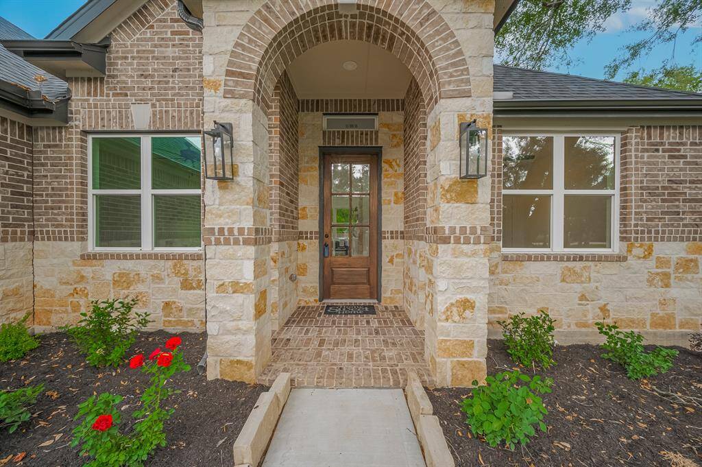 This inviting entrance features a charming brick and stone facade with an arched doorway, complemented by elegant outdoor lighting. Lush landscaping with blooming flowers adds a touch of color and warmth.
