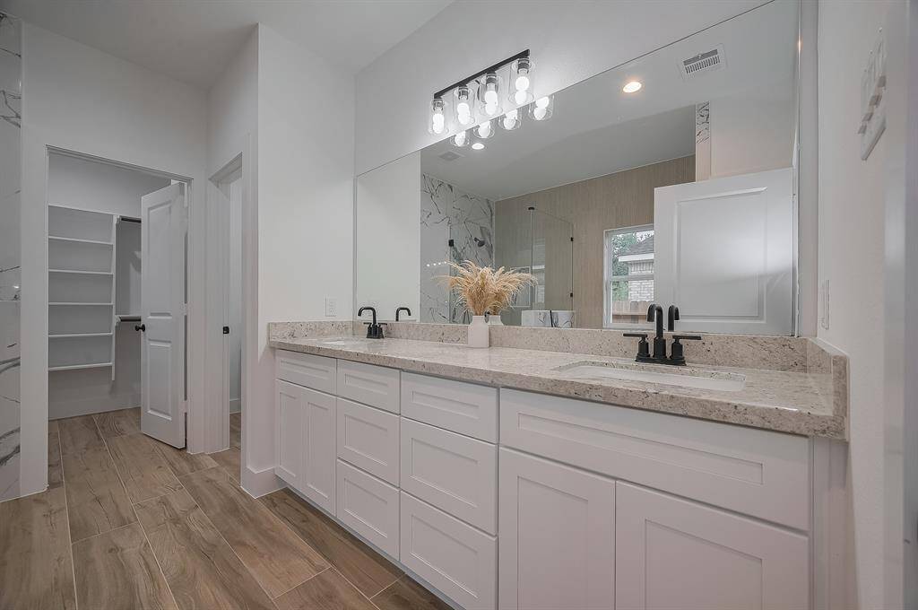 This modern bathroom features a spacious double vanity with granite countertops, sleek black fixtures, and ample cabinet space. It includes a large frameless mirror, wood-look tile flooring, and access to a walk-in closet with shelving.
