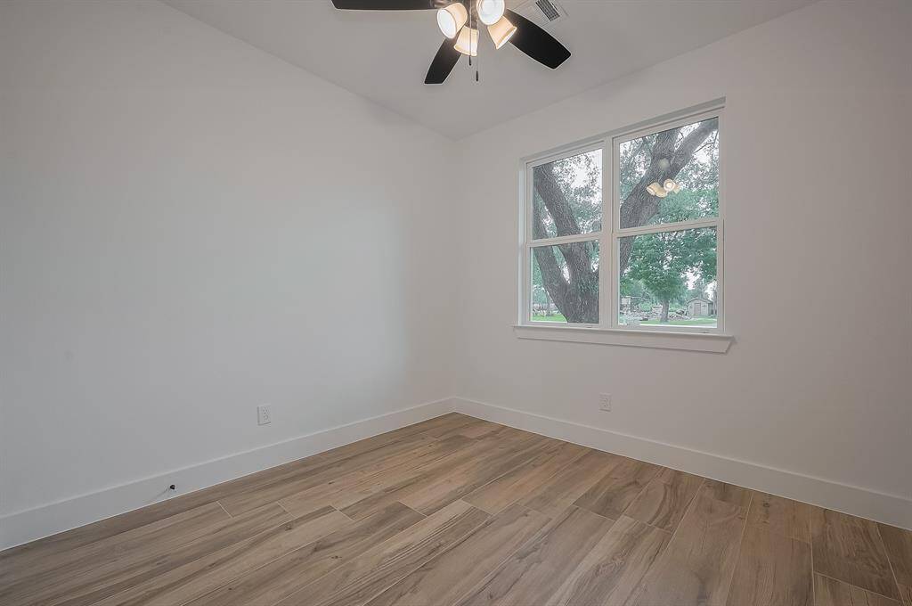 This room features fresh white walls, wood-style flooring, a ceiling fan with lights, and a large window offering views of lush greenery, creating a bright and inviting space.