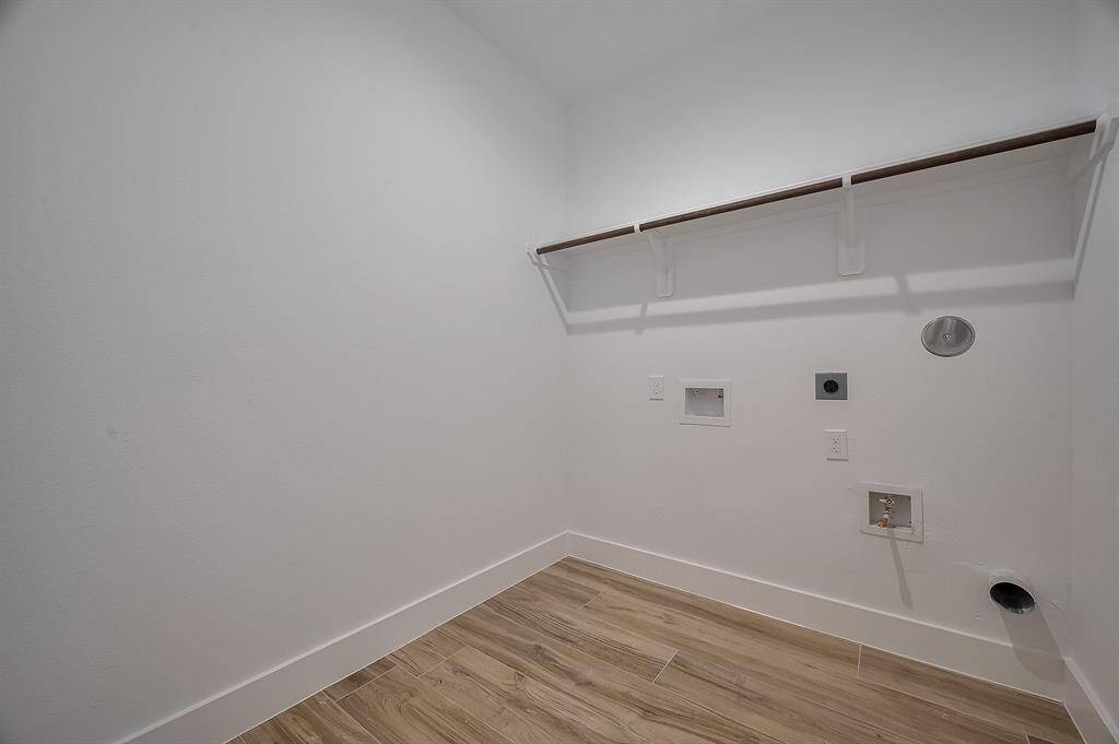 This is a utility room with plumbing and electrical connections for a washer and dryer. It features a wooden shelf and light-colored tile flooring, offering a clean and functional space.