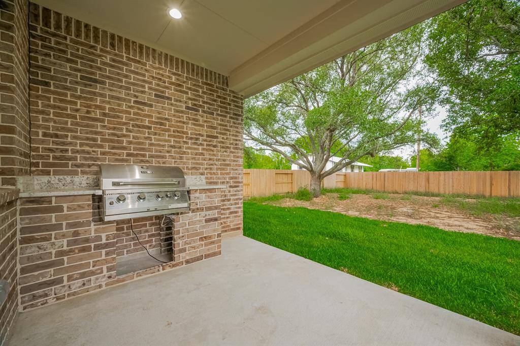 This photo shows a covered patio area with a built-in stainless steel grill set against a brick wall. The space opens up to a fenced backyard with green grass and a large tree, offering a perfect spot for outdoor entertaining.