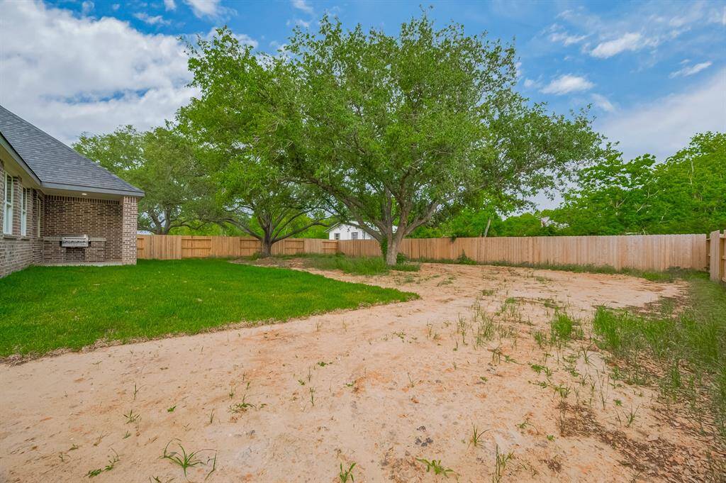 Spacious backyard featuring a blend of grass and sandy soil, bordered by a wooden fence. The area includes mature trees providing shade, ideal for outdoor activities or landscaping opportunities.