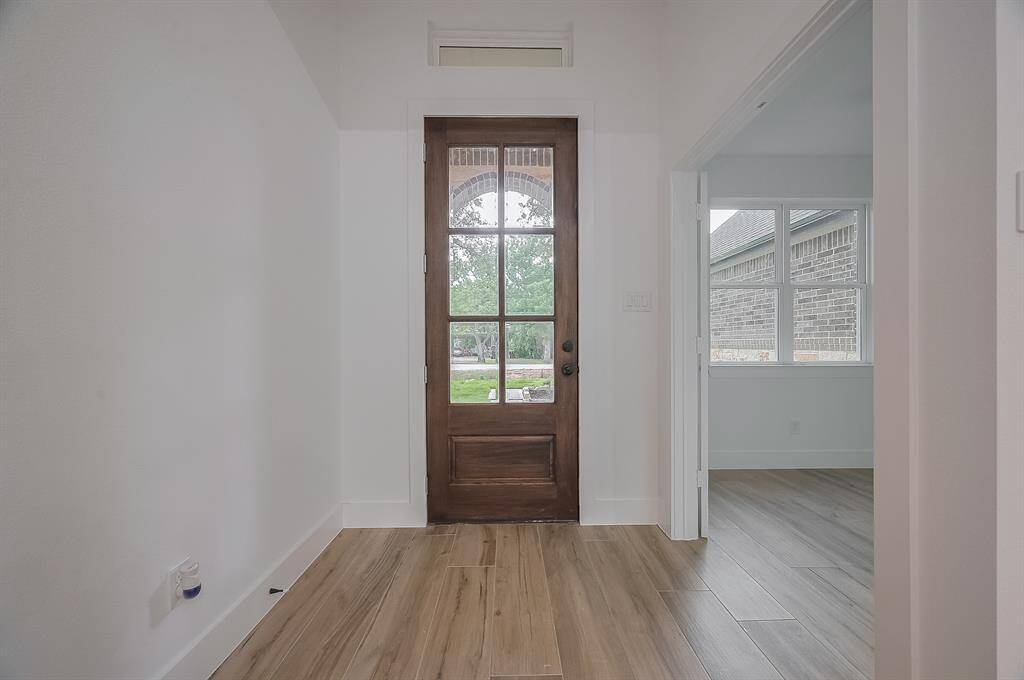 This entryway features a wooden front door with glass panels, allowing for natural light. The space has light wood flooring and neutral walls, offering a bright and welcoming atmosphere. An adjacent room with large windows is visible, enhancing the open feel.