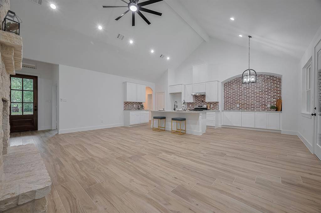 This spacious open-concept kitchen and living area features high vaulted ceilings, modern light fixtures, and a stylish blend of white cabinetry with brick accents. The room is bright and airy, enhanced by natural wood flooring and large windows, creating a cozy yet elegant atmosphere.