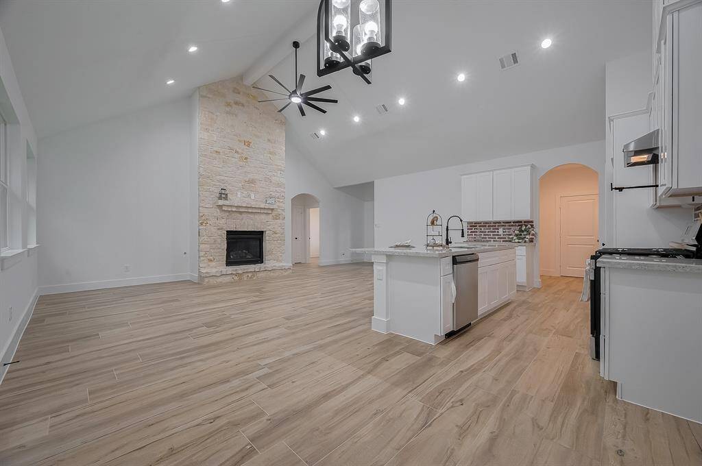 This spacious open-concept living area features a high vaulted ceiling, modern lighting, and a cozy stone fireplace. The kitchen boasts a large island, sleek cabinetry, and stainless steel appliances, perfect for entertaining. Warm wood-look flooring adds a touch of elegance.