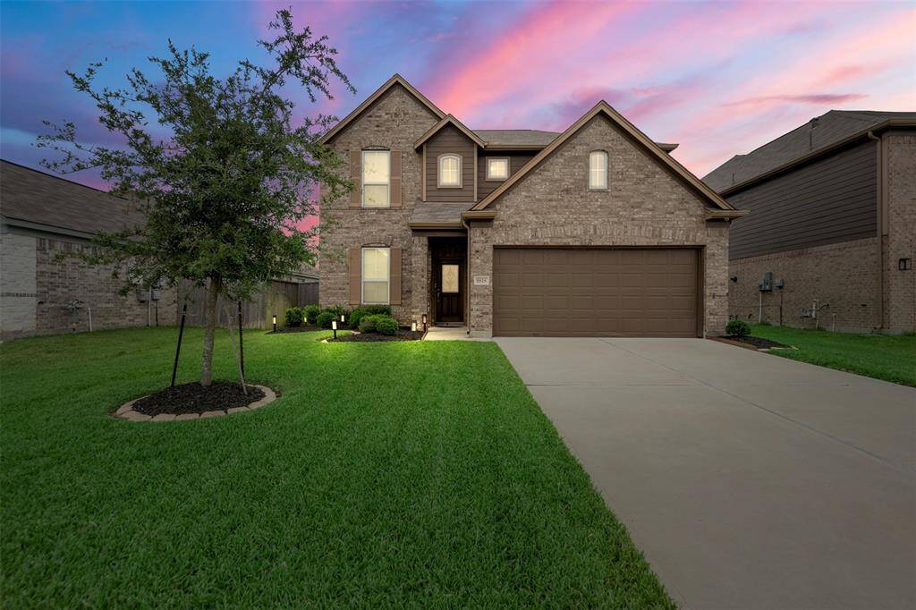 This photo showcases a two-story brick home with a neatly maintained front lawn and a young tree. The house features a two-car garage and is set against a vibrant sunset sky, creating a warm and inviting atmosphere.