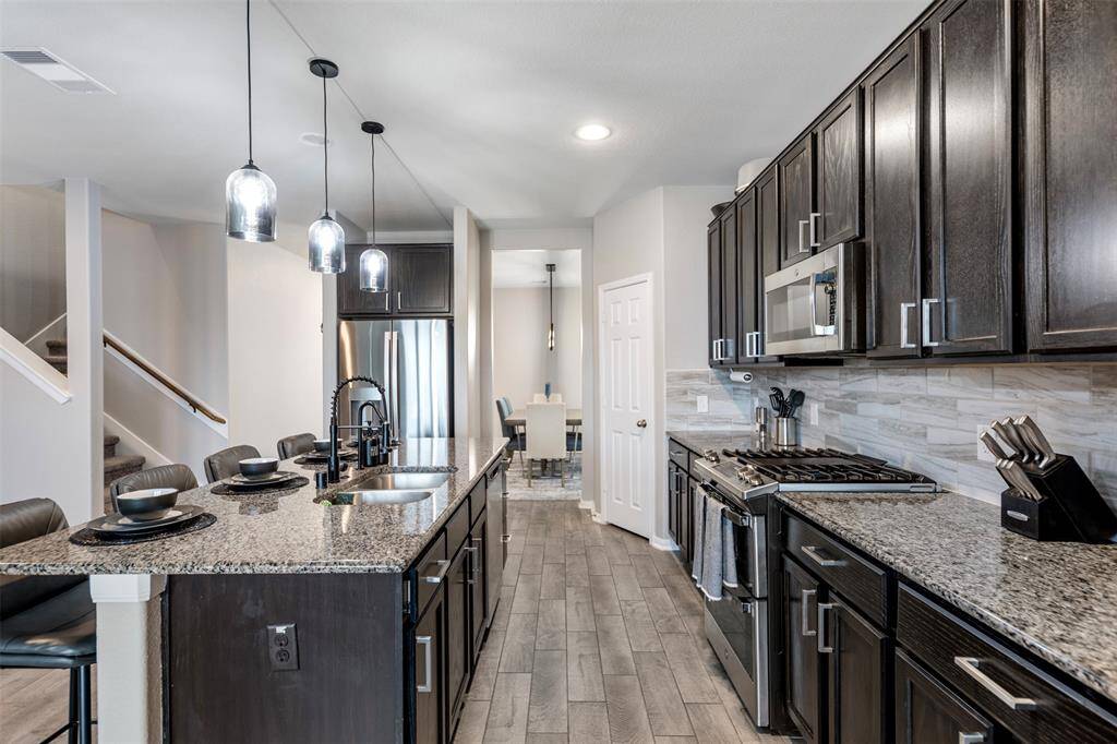 Modern kitchen with dark cabinetry, granite countertops, and stainless steel appliances. Features an island with seating, pendant lighting, and a view into the dining area. Neutral tones and wood-look tile flooring create a sleek, contemporary feel.