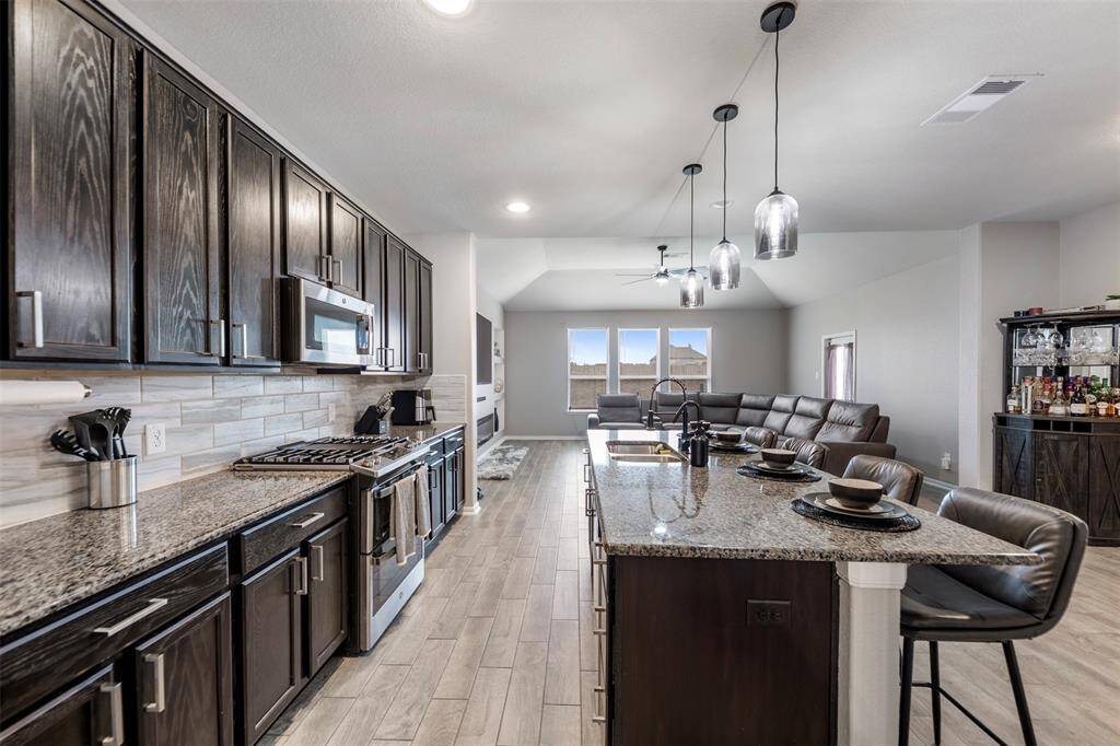 This photo showcases a spacious, modern open-concept kitchen and living area. The kitchen features dark wood cabinetry, granite countertops, a large island with seating, and stainless steel appliances. The adjoining living space is bright with natural light from multiple windows, offering a comfortable seating arrangement.