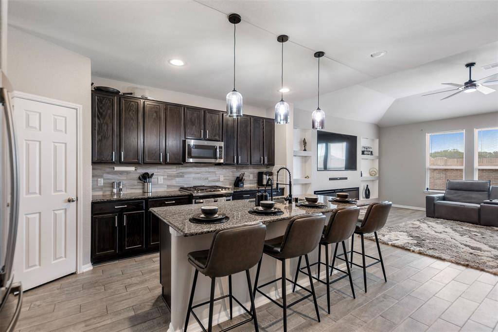 This modern open-concept kitchen and living area features dark wood cabinetry, a large island with seating for four, pendant lighting, and a cozy living space with a mounted TV and large windows for natural light.