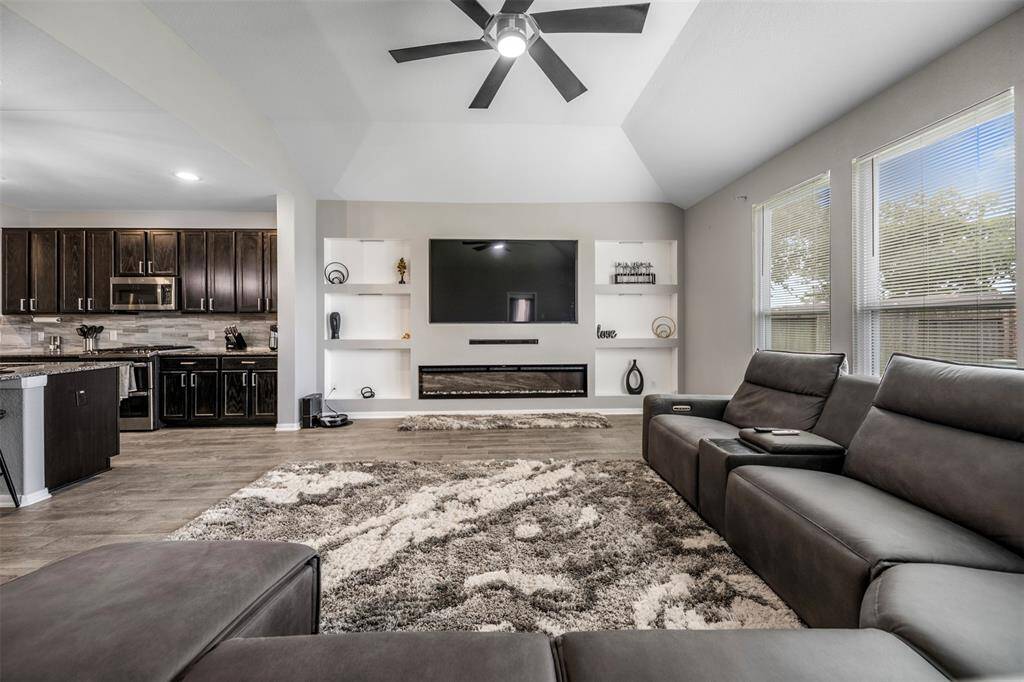 This spacious living room features modern gray seating, a large wall-mounted TV, and an electric fireplace. It seamlessly connects to an open kitchen with dark cabinetry. Natural light pours in from the large windows, highlighting the stylish decor and cozy carpet.