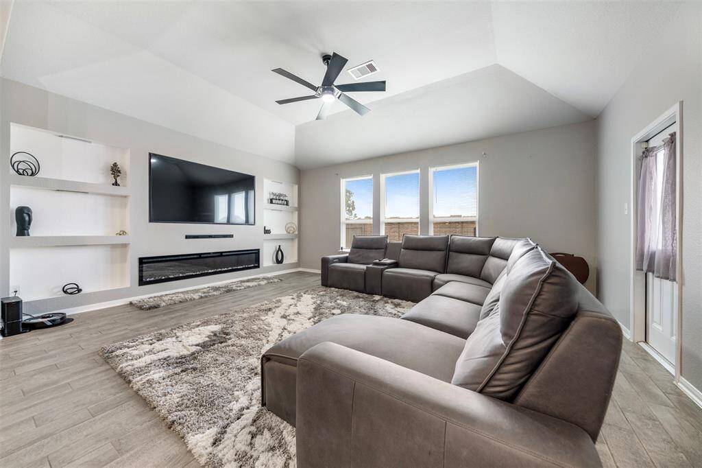This spacious living room features a modern design with a large sectional sofa and stylish wood-look flooring. It includes a mounted flat-screen TV and a sleek electric fireplace. Built-in shelving adds a decorative touch, while large windows let in natural light.