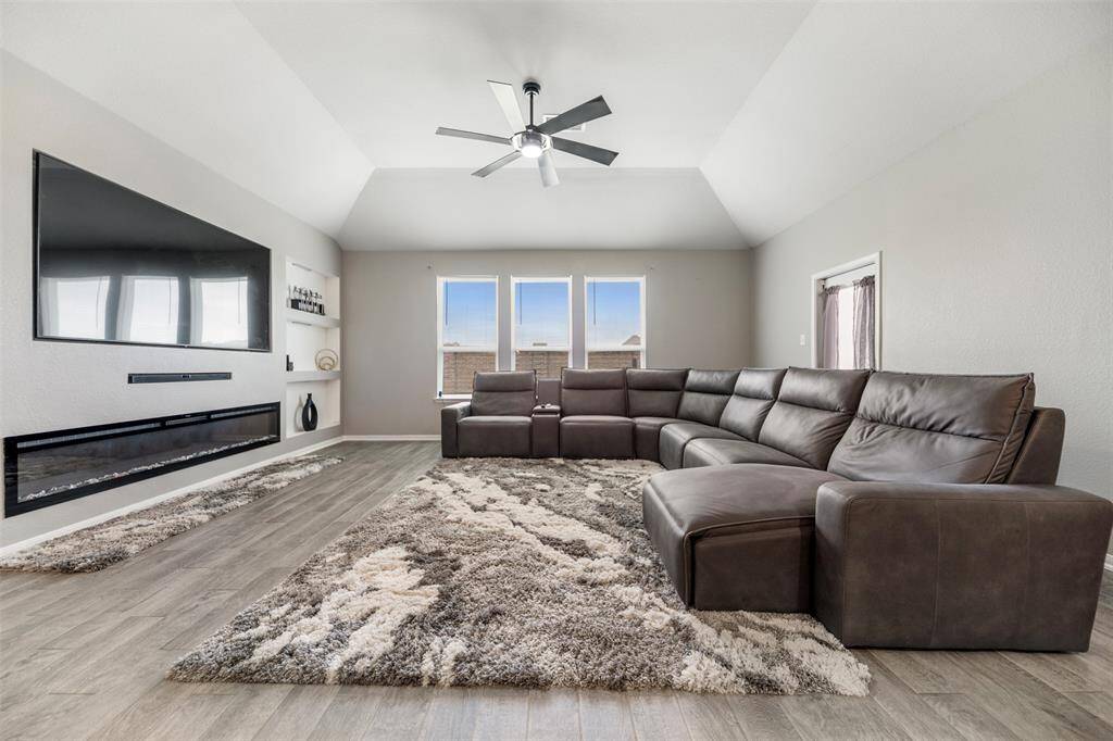 This spacious living room features a modern, open layout with a large sectional sofa, sleek wood flooring, and a cozy fireplace. A mounted flat-screen TV and ceiling fan complete the contemporary look, while natural light floods in through two windows. Ideal for relaxation and entertainment.