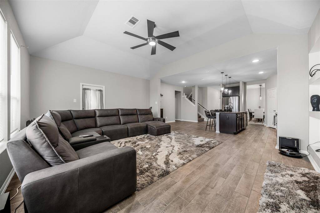 Spacious open-concept living area with high ceilings, featuring a large sectional sofa and modern ceiling fan. The room flows into a sleek kitchen with an island and dining area. Wood-look flooring and ample natural light create a warm, inviting atmosphere.