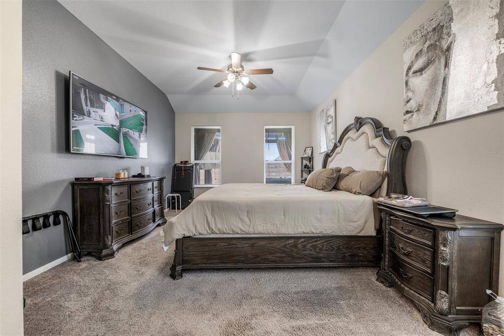 This spacious bedroom features a vaulted ceiling and a ceiling fan, providing a comfortable atmosphere. It includes a large bed with elegant wooden furniture, a wall-mounted TV, and natural light from two windows, offering a cozy and stylish retreat.