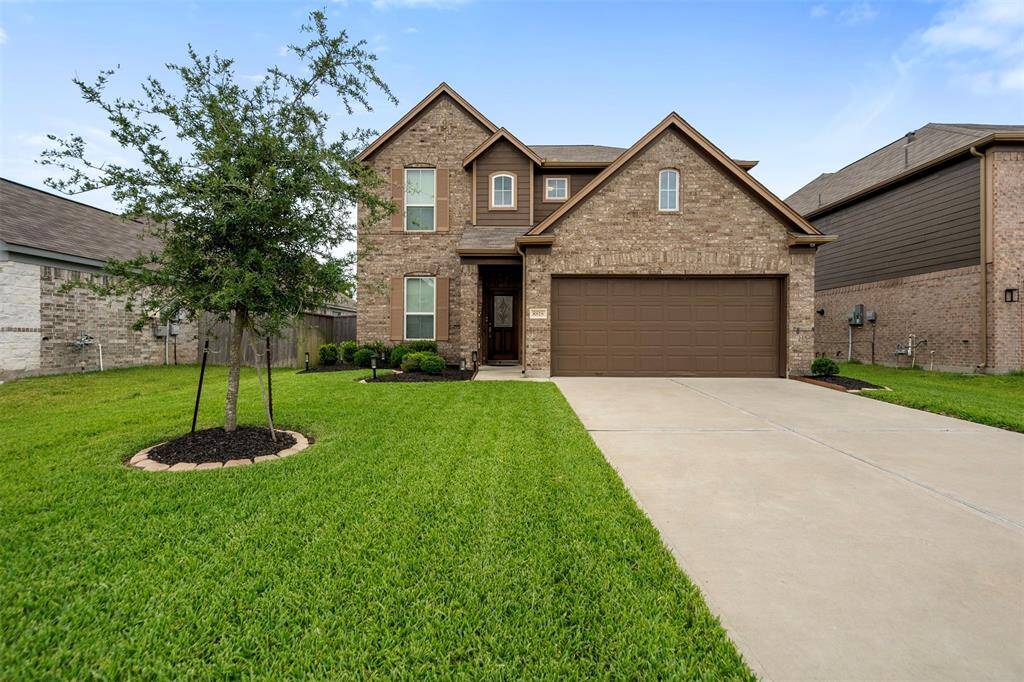 A charming two-story brick home with a well-maintained front yard and a spacious two-car garage. The driveway is wide, and there's a small tree adding greenery to the landscape. The house has a welcoming entrance and modern design.