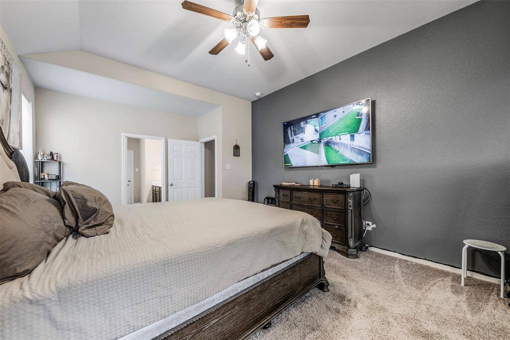 Spacious bedroom with neutral tones, featuring a ceiling fan and plush carpeting. A large TV is mounted on a dark accent wall, with a dresser beneath. The room has an en-suite bathroom and closet access, offering ample natural light and a cozy atmosphere.