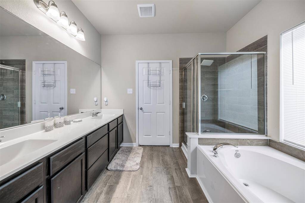 This bathroom features a modern design with a spacious double vanity, large mirror, and sleek lighting. It includes a glass-enclosed shower and a separate bathtub. The neutral color palette and wood-style flooring create a clean, elegant atmosphere.