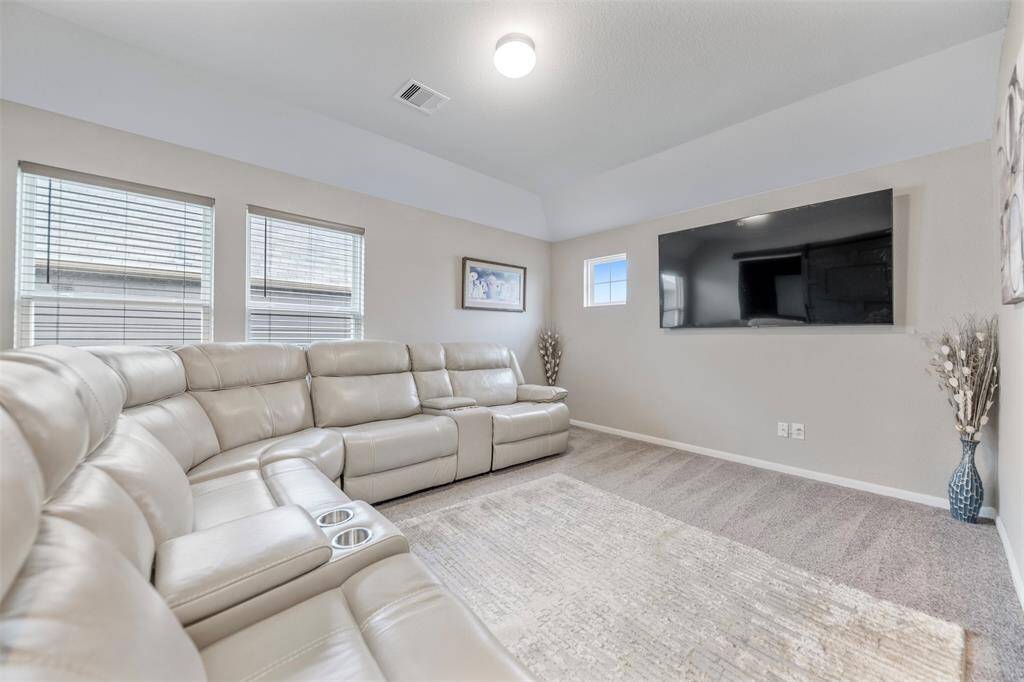 This cozy living room features a large sectional sofa, a mounted flat-screen TV, soft carpeting, and abundant natural light from multiple windows, creating a perfect setting for relaxation and entertainment.