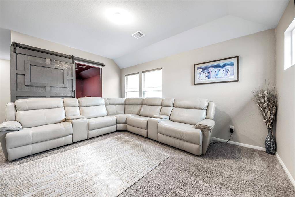 This cozy living room features a large L-shaped sectional sofa, perfect for relaxation. It has neutral walls, a soft carpet, and a stylish sliding barn door. Natural light fills the space through multiple windows, and tasteful decor adds a welcoming touch.