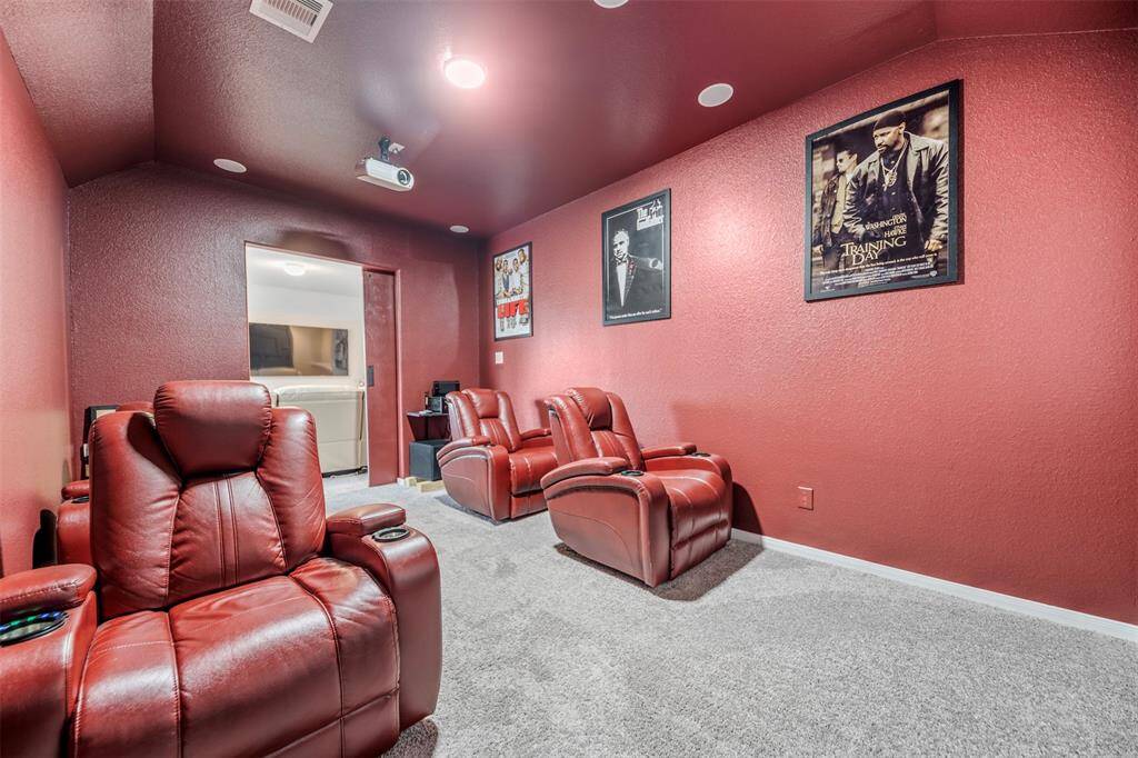 This photo shows a cozy home theater room with plush red recliner seats, maroon walls adorned with movie posters, and a ceiling-mounted projector. The space provides an intimate setting for enjoying films.