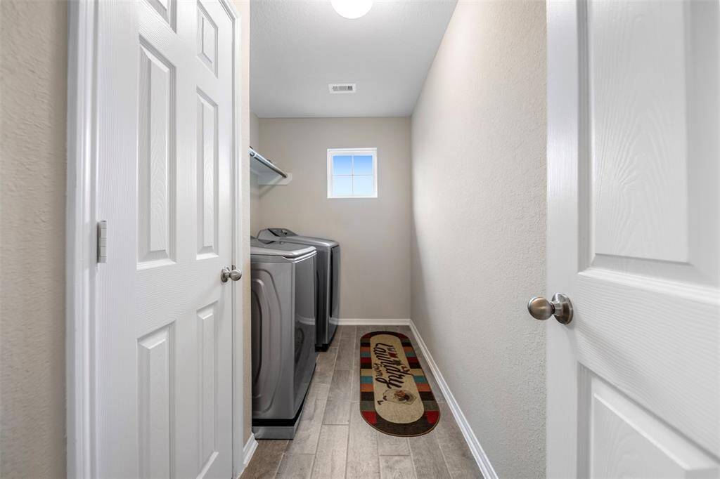 This photo shows a laundry room with a washer and dryer, neutral walls, and wooden flooring. It has a small window for natural light and is accessible through a white door.