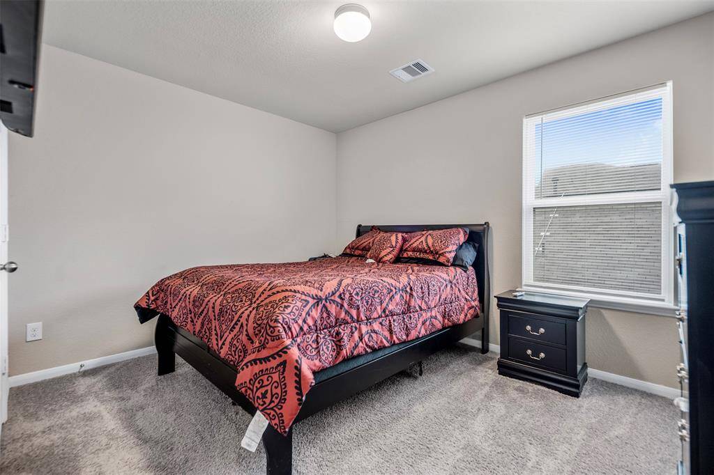 Cozy bedroom with soft carpet, neutral walls, and a large window offering natural light. It features a stylish black bed with a decorative red comforter and a matching nightstand.
