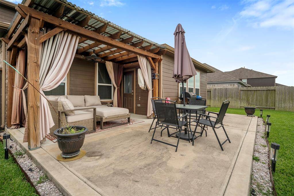 This photo showcases a cozy backyard patio with a wooden pergola featuring draped curtains, comfortable outdoor seating, a dining set with an umbrella, and a neatly maintained lawn, perfect for relaxing and entertaining.