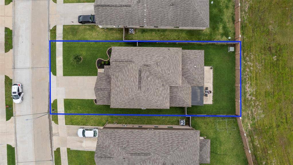 This aerial photo shows a single-family home with a neatly maintained yard, outlined in blue. The house has a driveway, a patio, and is situated in a suburban neighborhood with nearby homes.