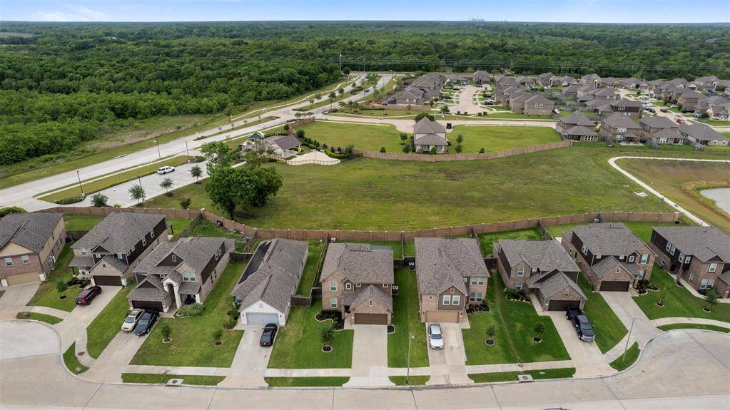 This aerial photo showcases a suburban neighborhood with modern, single-family homes, well-maintained lawns, and a backdrop of lush greenery. The area features a spacious layout with ample distance between homes and a nearby open green space, offering a peaceful, community-oriented environment.