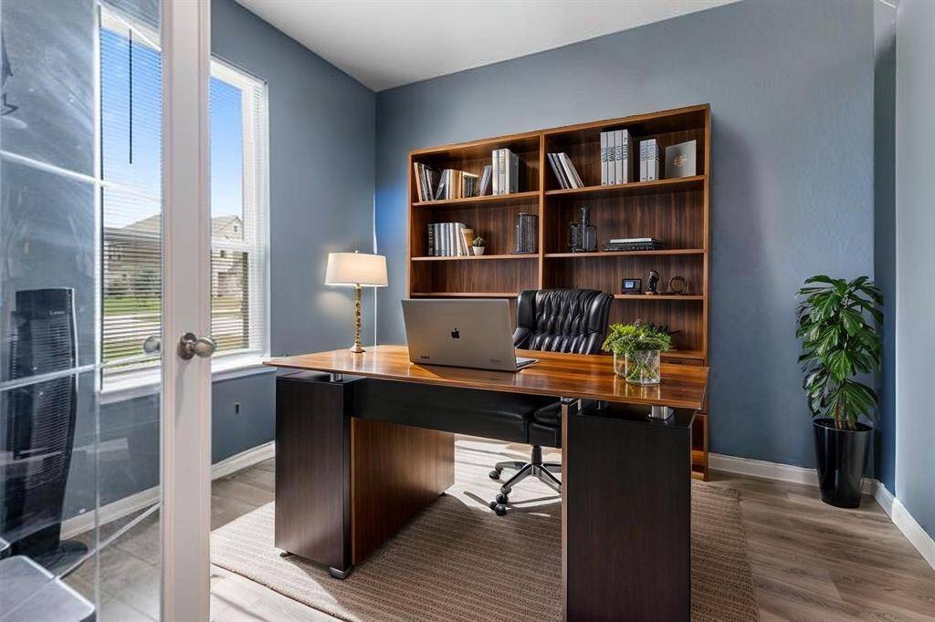 This home office features a modern desk with a laptop, a comfortable chair, and built-in wooden shelves filled with books and decor. Natural light streams in through a large window, highlighting the sleek blue walls and a potted plant, creating a cozy and productive workspace. Virtually staged.