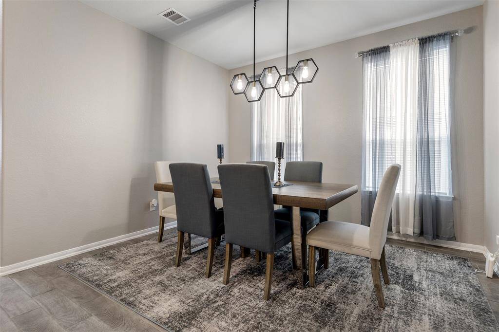 This dining room features a modern design with a stylish chandelier, neutral walls, and a cozy rug. It includes a wooden table with six upholstered chairs, creating a welcoming space for gatherings. Large windows allow natural light to brighten the room.