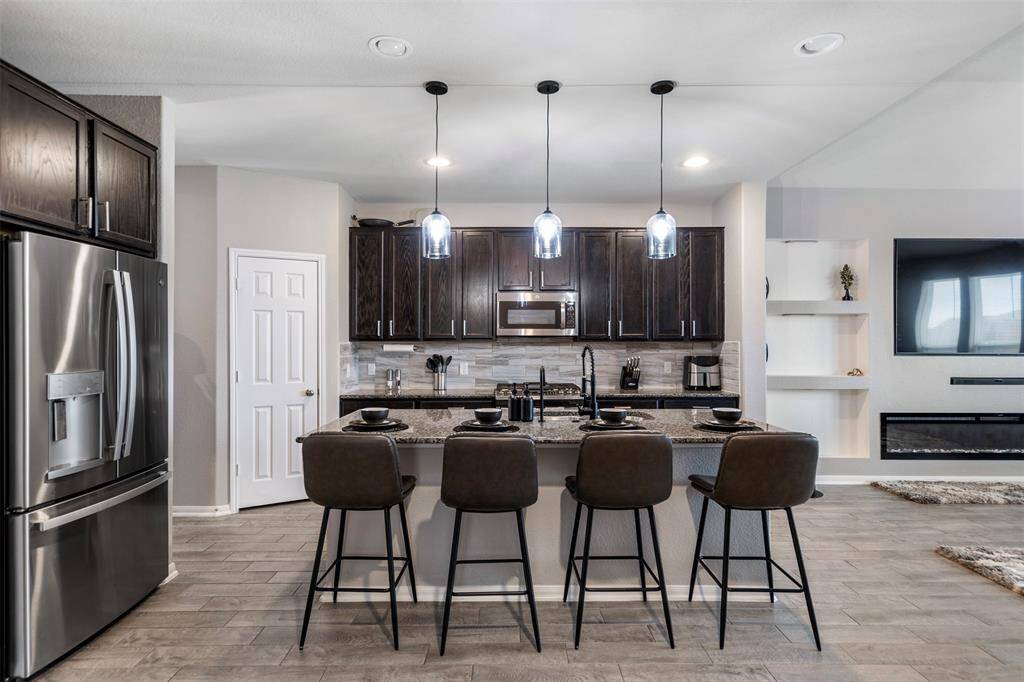 This modern kitchen features dark cabinetry, stainless steel appliances, and a stylish island with seating for four. Pendant lighting adds elegance, and the open layout connects to a cozy living area with a wall-mounted TV and fireplace.