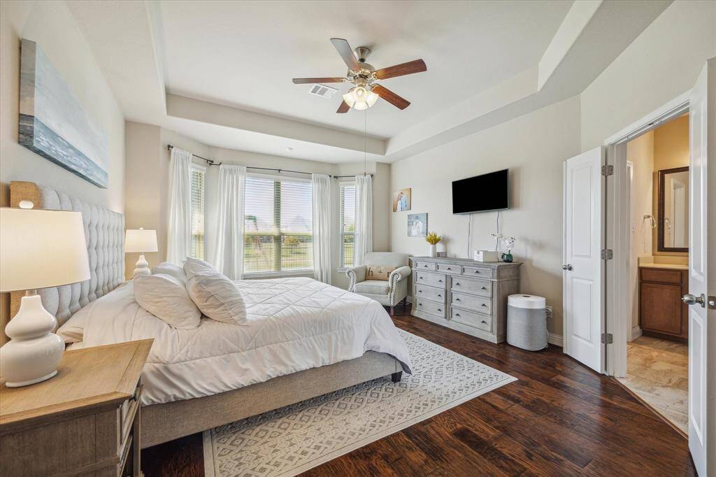 This inviting bedroom features a tray ceiling, hardwood floors, and large windows offering ample natural light. It includes a cozy seating area, modern ceiling fan, and en-suite bathroom, creating a comfortable and stylish retreat.