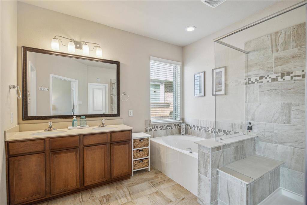 This primary bathroom features a double sink vanity with a large mirror, a spacious walk-in shower with glass doors, and a soaking tub. Natural light fills the room through a window, highlighting the elegant tile work and neutral tones.