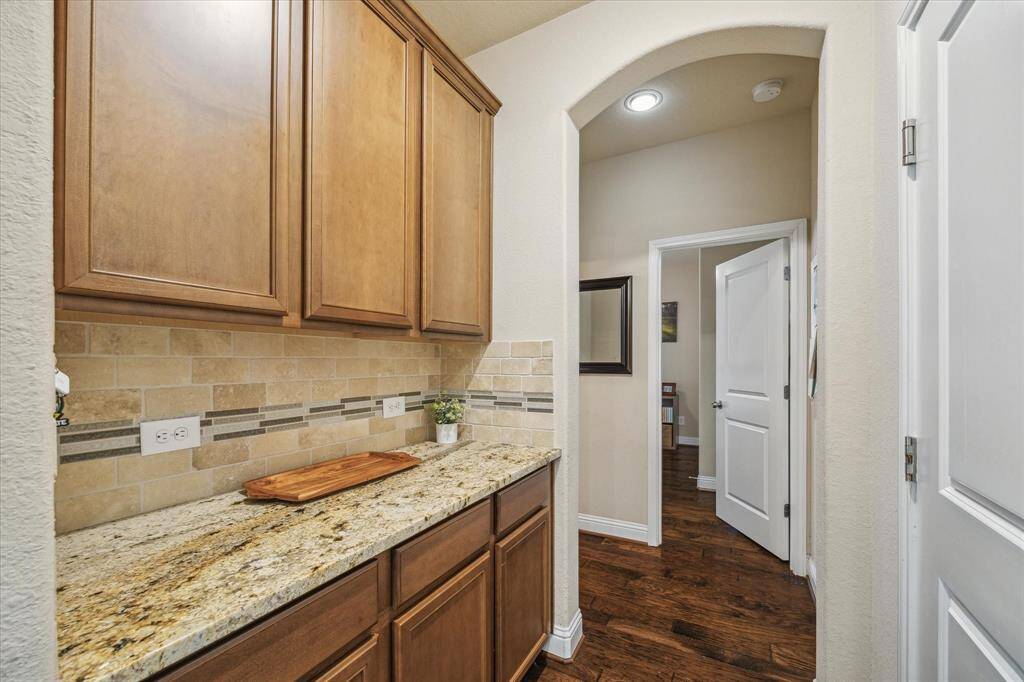 This photo showcases a hallway with warm wood cabinetry and granite countertops, ideal for extra storage, a coffee station, charging station for cell phones/ipads or spill over bar/china cabinet for kitchen items. It boasts an elegant tile backsplash, and the same high-end granite as the kitchen.  Conveniently located right inside the garage door and near the laundry room.  First floor secondary bedroom right down the hall.