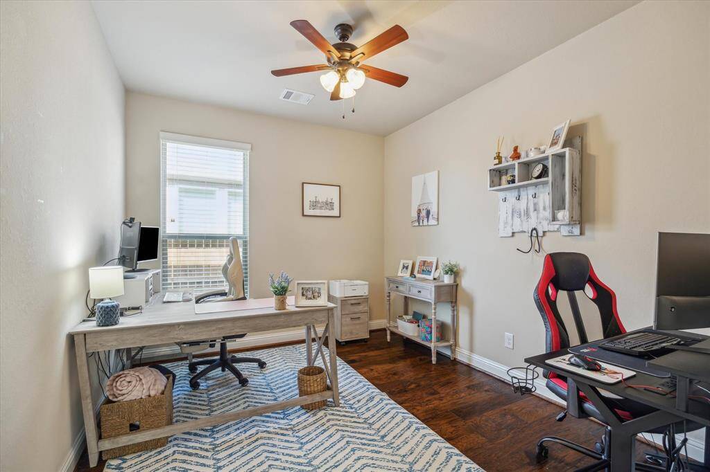 A bright home office with hardwood floors and neutral walls. It features a large window, a ceiling fan, and ample space for desks and shelving. Ideal for remote work or study.
