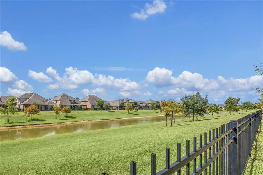 This photo features a serene neighborhood view with modern homes alongside a picturesque pond, lush green lawns, and a black metal fence in the foreground, under a clear blue sky.