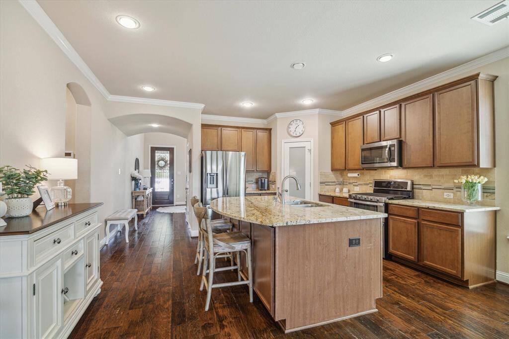 This kitchen features elegant wooden cabinetry, granite countertops, and stainless-steel appliances. The layout includes an island with seating, and the warm hardwood floors extend into the hallway, and throughout most of the first floor.