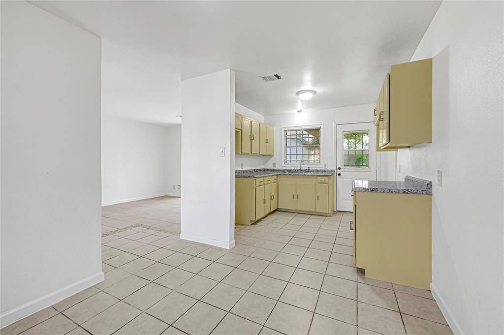 Bright kitchen with beige cabinets and granite countertops, featuring tiled flooring. Adjacent open area offers flexible living space. A window and glass door provide natural light and backyard access.