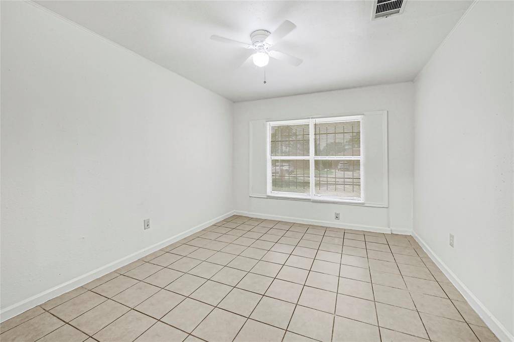 This secondary bedroom room features neutral-toned tiled flooring, a ceiling fan, and large windows with blinds, offering ample natural light. The space is versatile, ideal for a bedroom or home office.