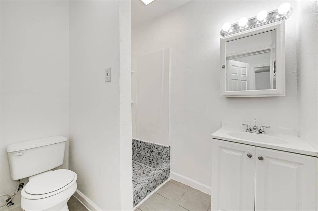 This bathroom features a minimalist design with white walls, a compact vanity with a mirror and bright lighting, a toilet, and a shower area with pebble tile detailing.