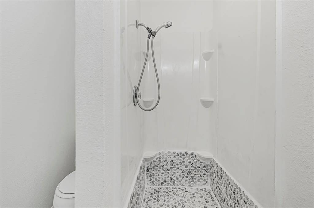 This bathroom features a modern shower with a handheld showerhead and stylish pebble-tiled flooring. Simple white walls create a clean, fresh look.