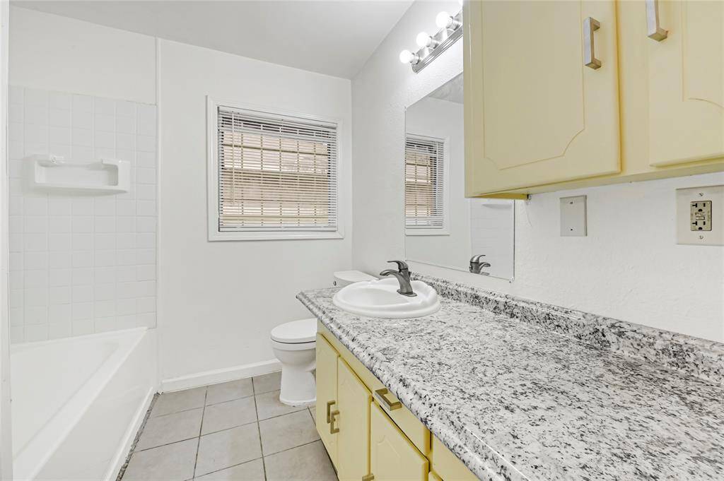 This bathroom features a sleek granite countertop, a large mirror, and ample storage with yellow cabinets. It includes a bathtub, neutral tiles, and a window for natural light, creating a bright and functional space.