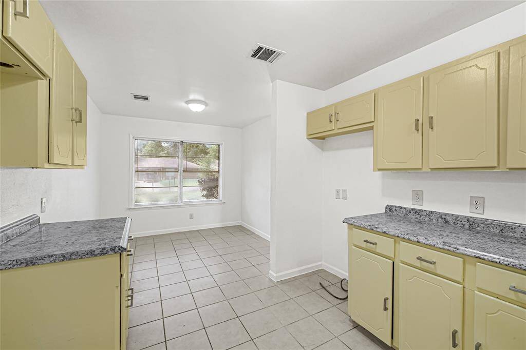 This kitchen features light yellow cabinets with gray countertops, a tiled floor, and a large window that allows natural light. The space is open, providing room for dining or additional kitchen storage.
