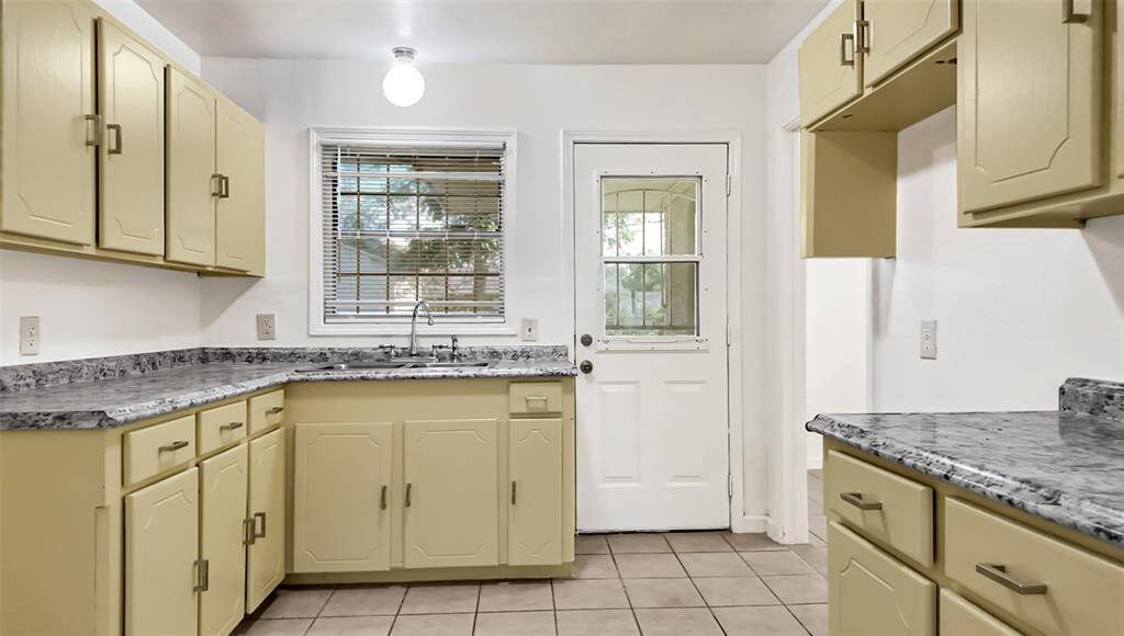 This kitchen features vintage-style yellow cabinetry with ample storage, complemented by gray marble-look countertops. A window above the sink and a door with glass panes provide natural light, enhancing the bright and welcoming atmosphere. Tile flooring adds durability and ease of maintenance.