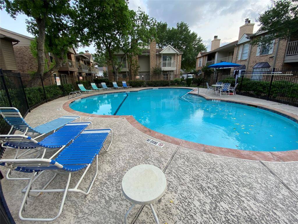 This photo features a well-maintained residential pool area, surrounded by trees and brick buildings. There are several lounge chairs and a small table on the deck, providing a relaxing space for residents.