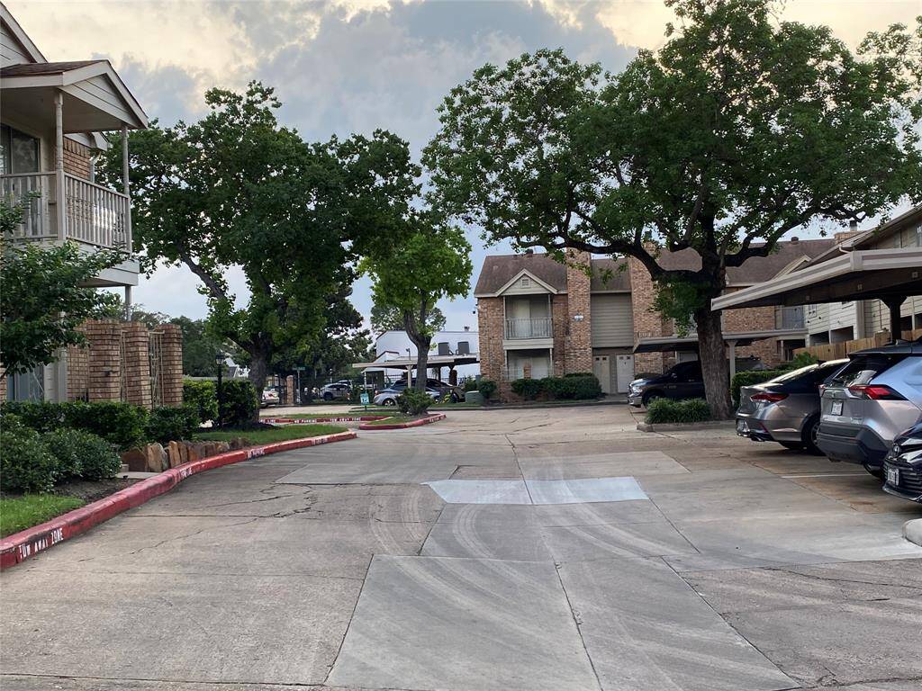 This photo shows a quiet residential area with brick buildings, mature trees, and well-maintained landscaping. There are parked cars under a covered area, creating a cozy neighborhood feel.