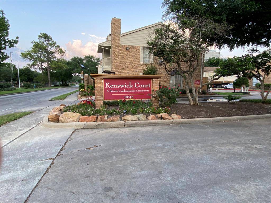 The photo shows the entrance to Kenswick Court, a private condominium community. It features a well-maintained sign surrounded by landscaped greenery and rocks, with a paved driveway and trees in the background, indicating a peaceful residential area.