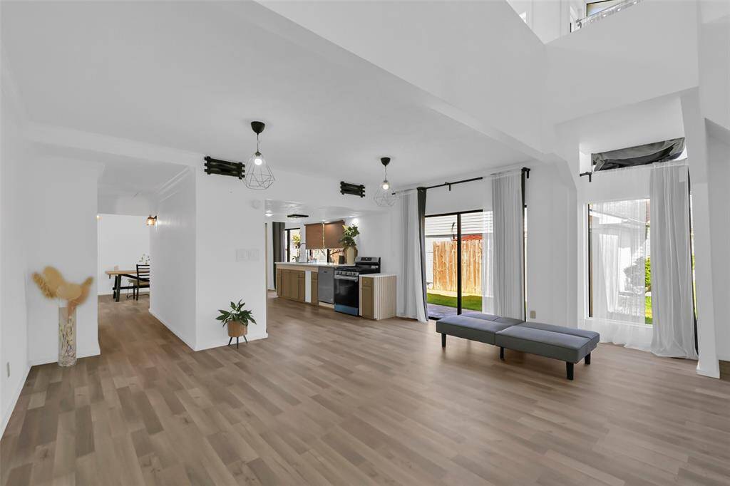 Bright and spacious open-concept living area with modern light fixtures and large windows. Light flooring throughout enhances the airy feel, while sliding doors provide easy access to the backyard.