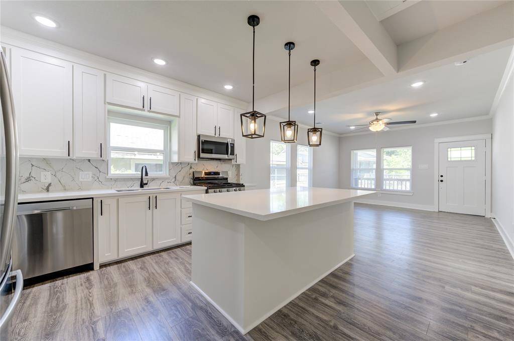 This photo showcases a modern, open-concept kitchen and living area. The kitchen features sleek white cabinetry, a marble backsplash, and stainless steel appliances. An island with pendant lighting provides a stylish focal point. The space has wood-look flooring and ample natural light, creating a bright and inviting atmosphere.